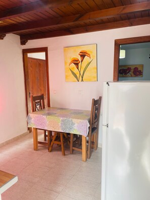 A dining area with a table covered by a floral tablecloth, wooden chairs, a white refrigerator, and a wooden door. - Margarita Penthouse Conil Center – Balcony & Air Conditioning (Conil de la Frontera)