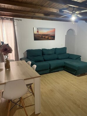 Ein Wohnzimmer mit einem grünen Ecksofa, einem Esstisch mit Stühlen, einer Vase mit Blumen und einem Gemälde eines Leuchtturms an der Wand.