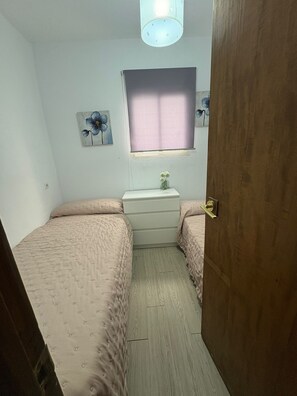 A small bedroom with two beds, a dresser, and a window with a curtain. - Casa Margarita Conil – Spacious, 3 Bedrooms, Historic Center (Conil de la Frontera)