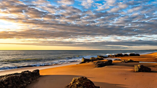 Uma praia com céu claro, ondas e afloramentos rochosos.