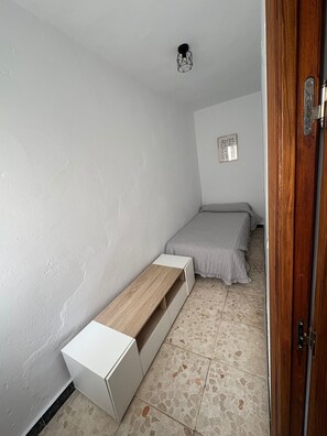 Ein kleiner, schmaler Raum mit einem Bett, einer Holzbank und einer Tür.