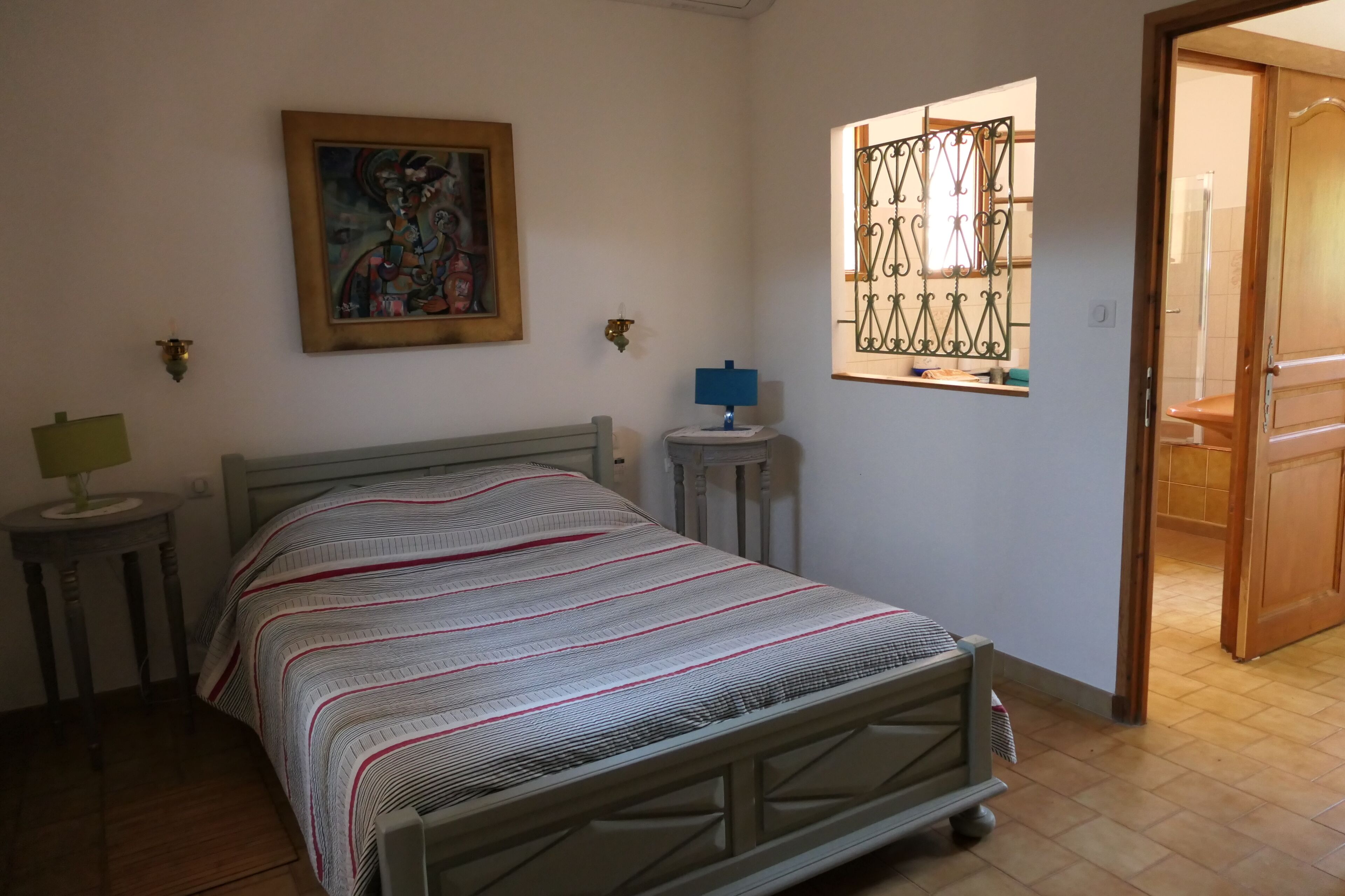 Een slaapkamer met een bed, een nachtkastje, een lamp, een aan de muur hangend schilderij en een raam met een metalen traliewerk.