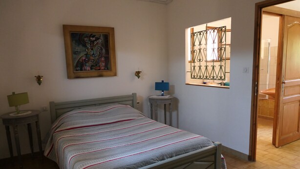 A bedroom with a bed, a bedside table, a lamp, a wall-mounted picture, and a window with a metal grille. - Provencal-style farmhouse with charm, swimming pool, and landscaped grounds on 6,200 m² (Draguignan)