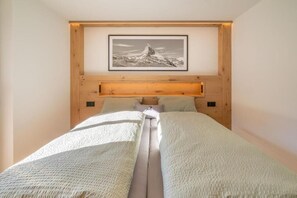 A bedroom with a bed centered between two wooden headboards, a framed picture above the bed, and a light-colored wall. - Apartment Bergliebe (Zermatt)