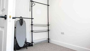 Ein Zimmer mit einer schwarzen Metallregaleinheit an einer weißen Wand, darin ein Staubsauger und ein graues Bügelbrett.