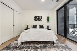Ein Schlafzimmer mit einem großen Bett, einem Nachttisch, einer Wandlampe, einer hängenden Pflanze und einer Glastür, die zu einem Außenbereich führt.
