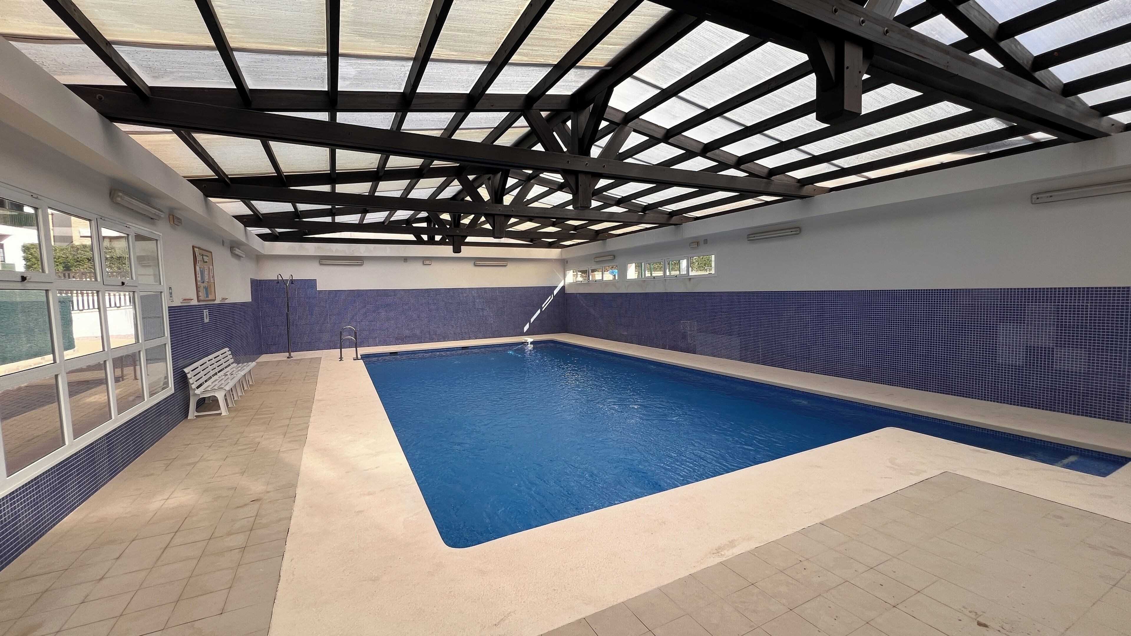 Une piscine intérieure avec un sol et des murs carrelés, un plafond doté d’une lucarne et un banc le long du bord.