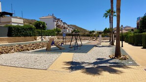 A playground with swings and a slide, surrounded by a paved area with palm trees and a stone wall. - Quiet 2-bedroom chalet in Vera Playa | Pool & Near the beach (Vera)