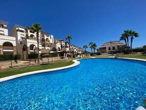 Une grande piscine entourée d’immeubles à plusieurs étages dotés de balcons, de palmiers et surplombant un ciel bleu azur.
