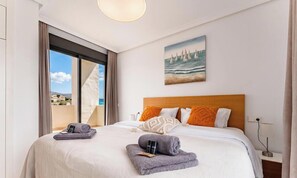 Quarto de hotel com uma cama grande, uma pintura de veleiros e vista para o mar pela janela.