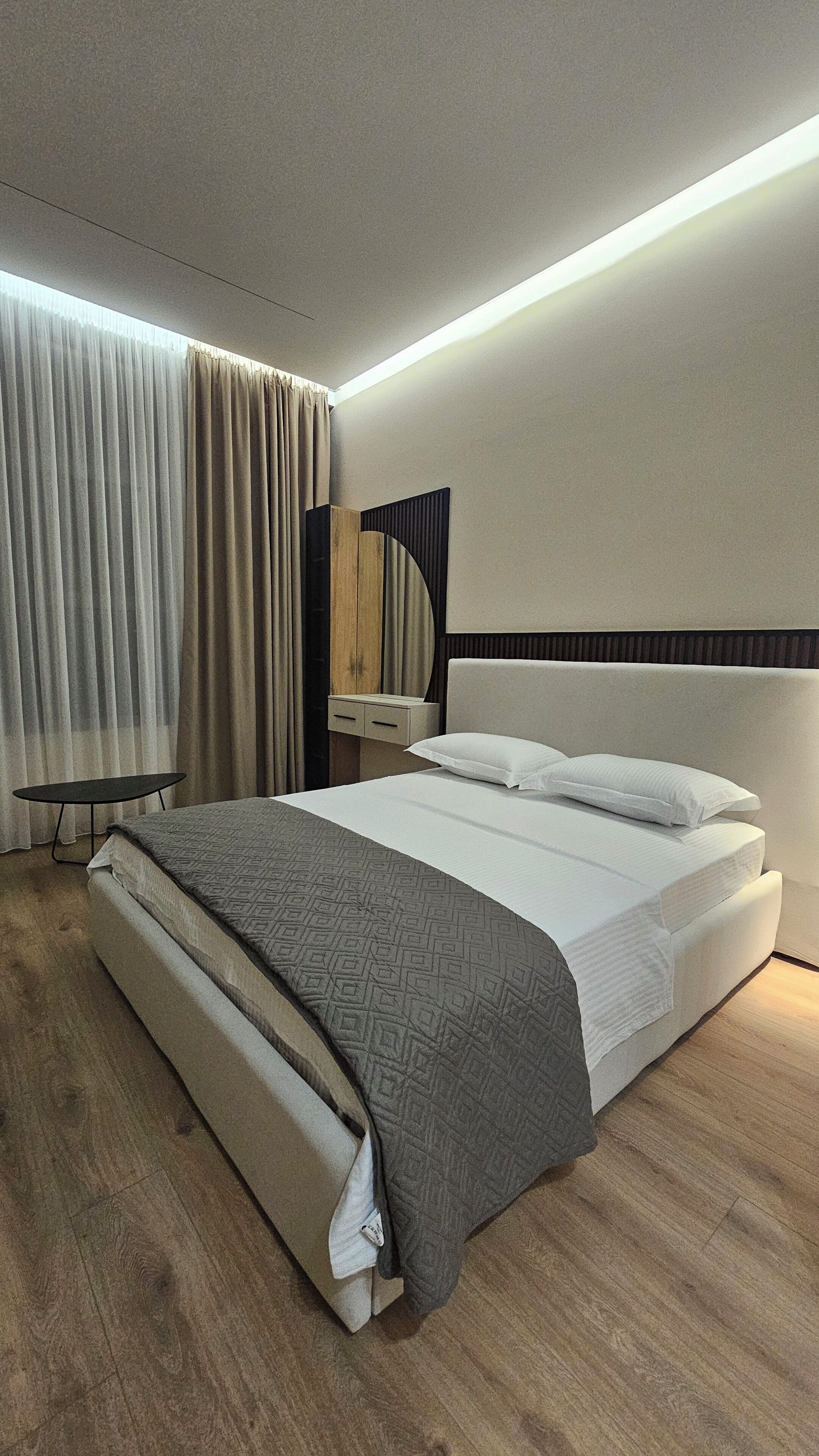 A modern hotel room with a large bed, a bedside table, a mirror, and a window with curtains.