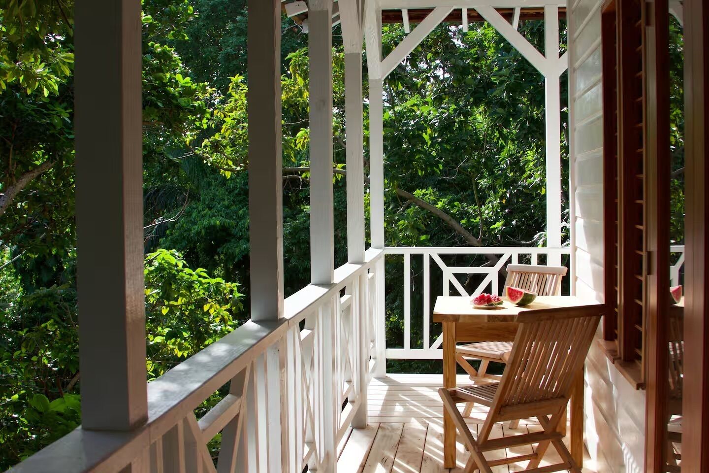 A wooden porch with a table and chair, surrounded by white railings and lush greenery.