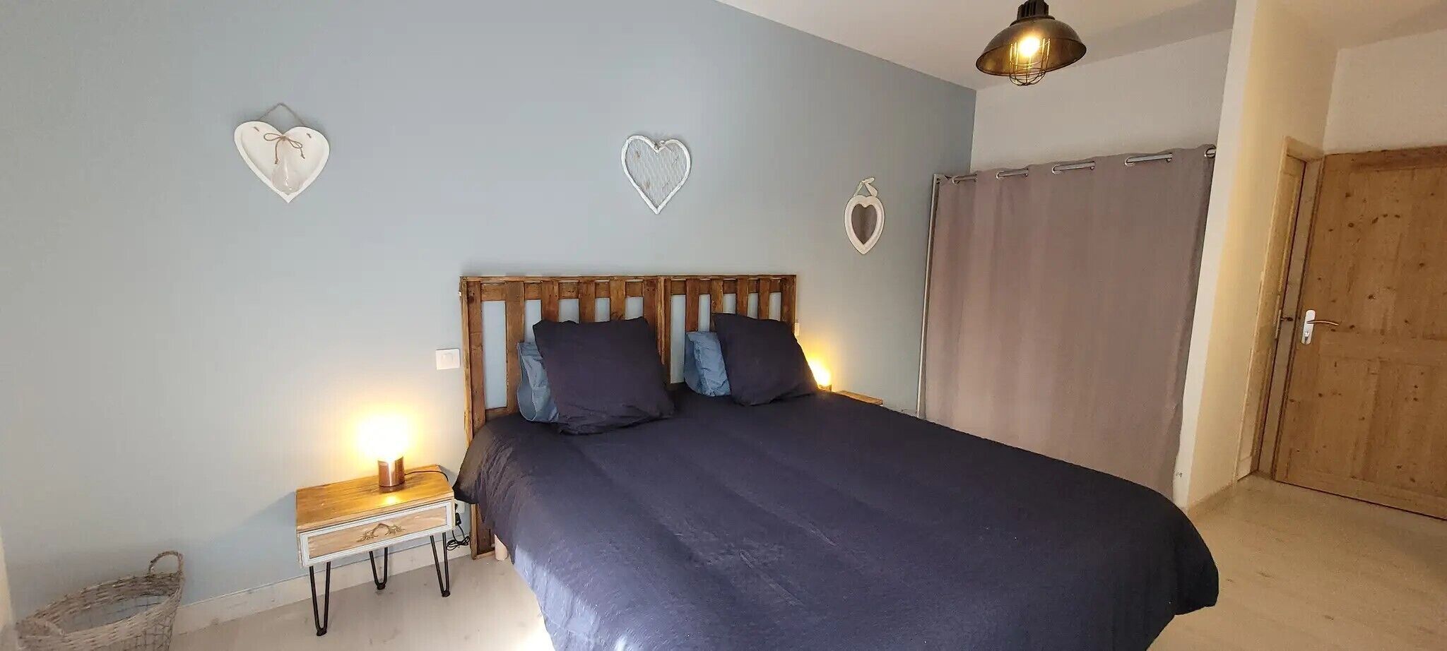 A bedroom with a wooden headboard, a bed with dark bedding, a bedside table with a lamp, and heart-shaped wall decorations.