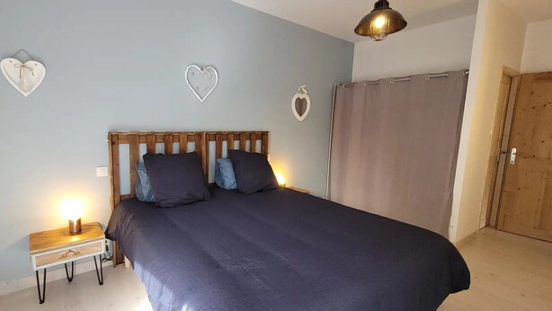 A bedroom with a wooden headboard, a bed with dark bedding, a bedside table with a lamp, and heart-shaped wall decorations. - “Cézallier Room” Guest Suite with Private Terrace and Jacuzzi (Soulages)