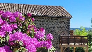 A stone building with a tiled roof, a wooden deck, and blooming pink flowers in the foreground. - “Cézallier Room” Guest Suite with Private Terrace and Jacuzzi (Soulages)