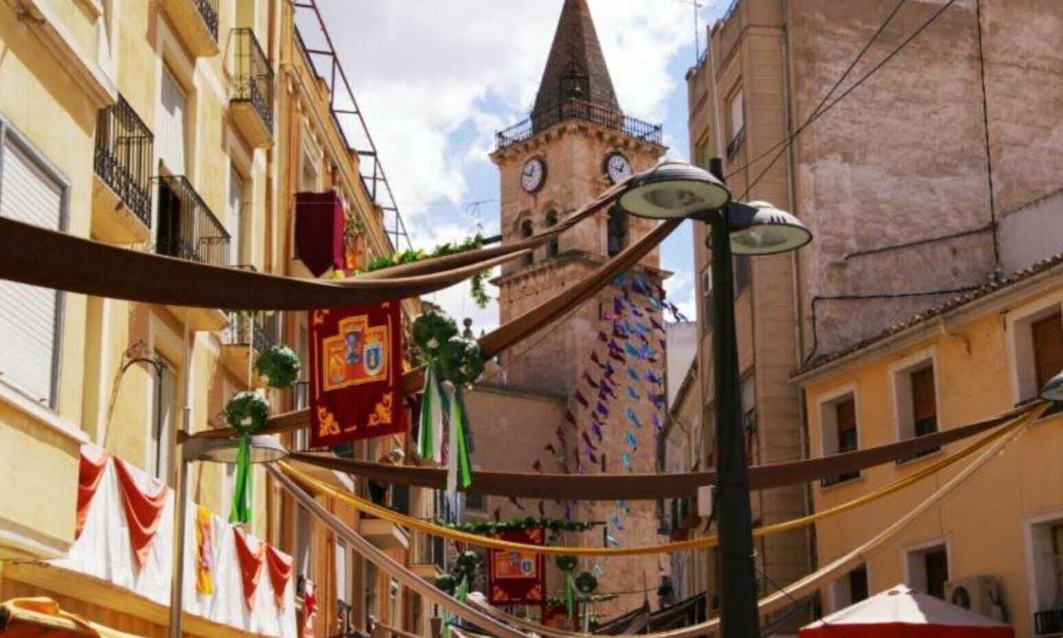 A narrow street with buildings, a tower with clocks, and hanging decorations.