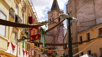 A narrow street with buildings, a tower with clocks, and hanging decorations.
