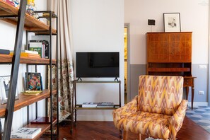 Ein Wohnzimmer mit Fernseher, einem gemusterten Sessel, einer Holzkommode und einem Bücherregal mit Büchern und Dekoration.