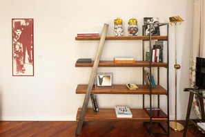 Ein Zimmer mit Holzboden, einem Bücherregal mit verschiedenen Büchern, einem gerahmten Bild an der Wand und einer Stehlampe.
