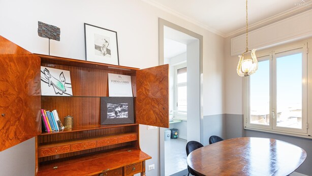 Un coin repas avec une table ronde en bois et des chaises, une bibliothèque contenant divers objets et une fenêtre laissant entrer la lumière naturelle.