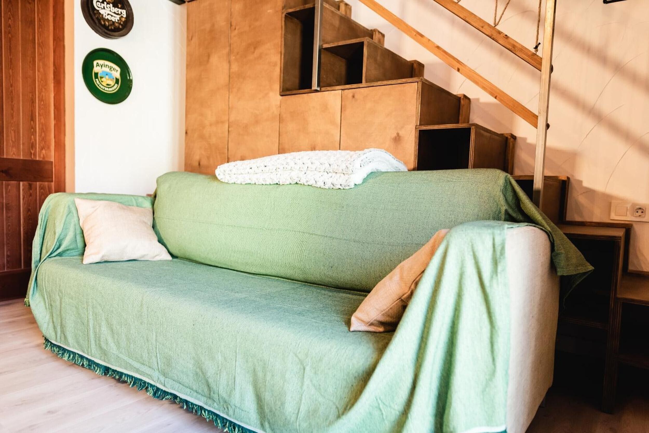 A green sofa with a white throw blanket and pillows, wooden walls, and a staircase with a wooden railing.