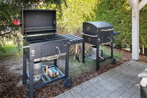 Una griglia per barbecue con un bruciatore laterale e un affumicatore, entrambi su supporti con ruote, immersi in un giardino con vialetto in mattoni.