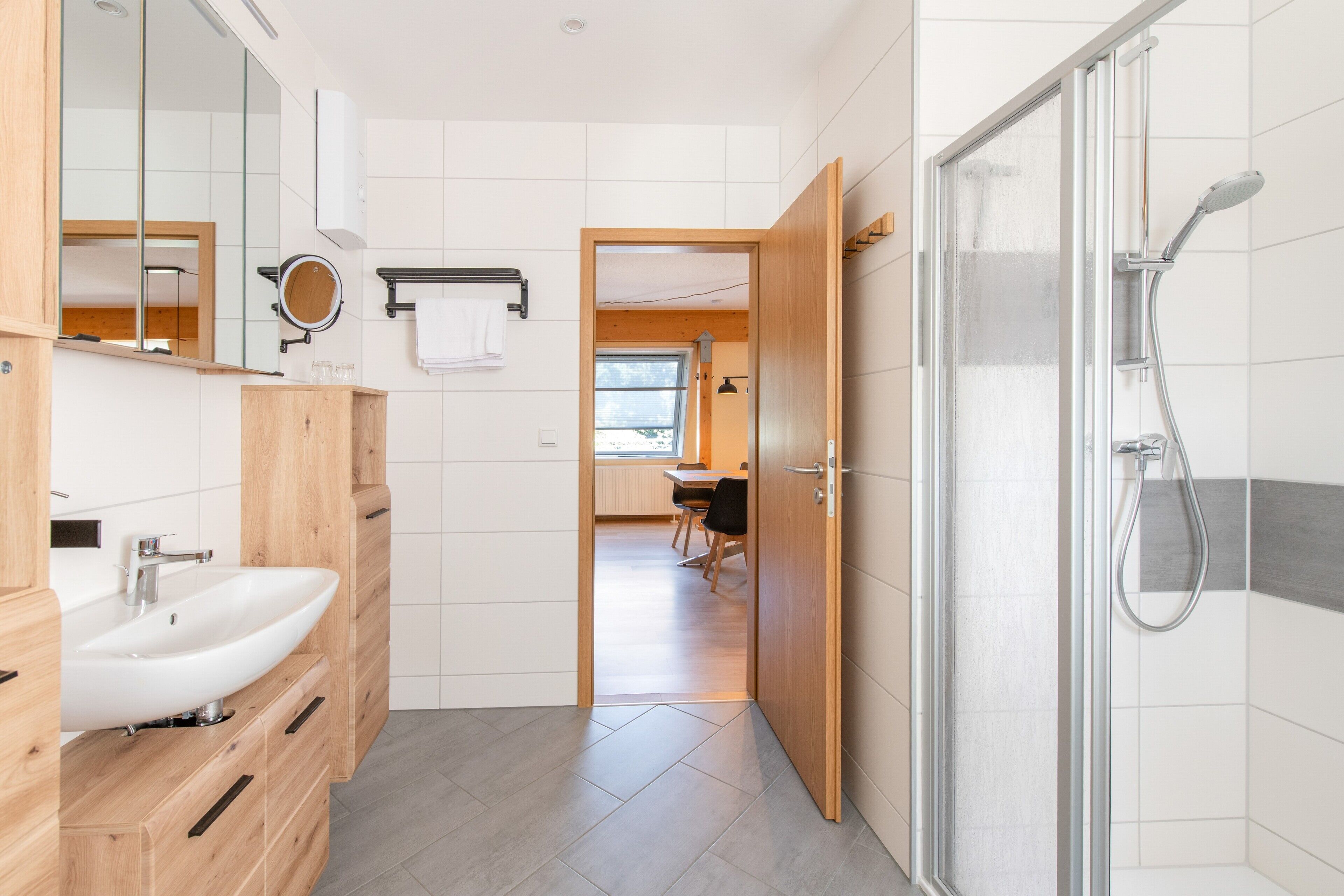 Ein modernes Badezimmer mit Dusche, Waschbecken und Spiegel. Es gibt einen Holzkästen und einen Handtuchhalter.