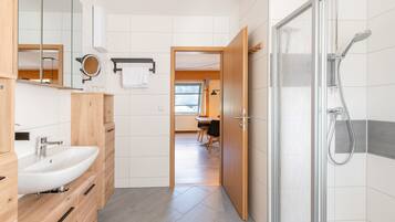 Ein modernes Badezimmer mit Dusche, Waschbecken und Spiegel. Es gibt einen Holzkästen und einen Handtuchhalter.