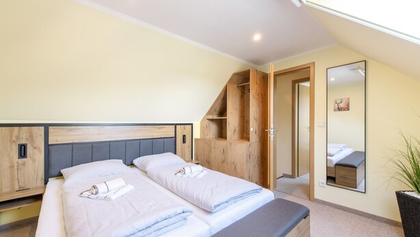 Ein Doppelbett mit grauem Kopfteil, ein Holzschrank, ein Spiegel und eine Pflanze.