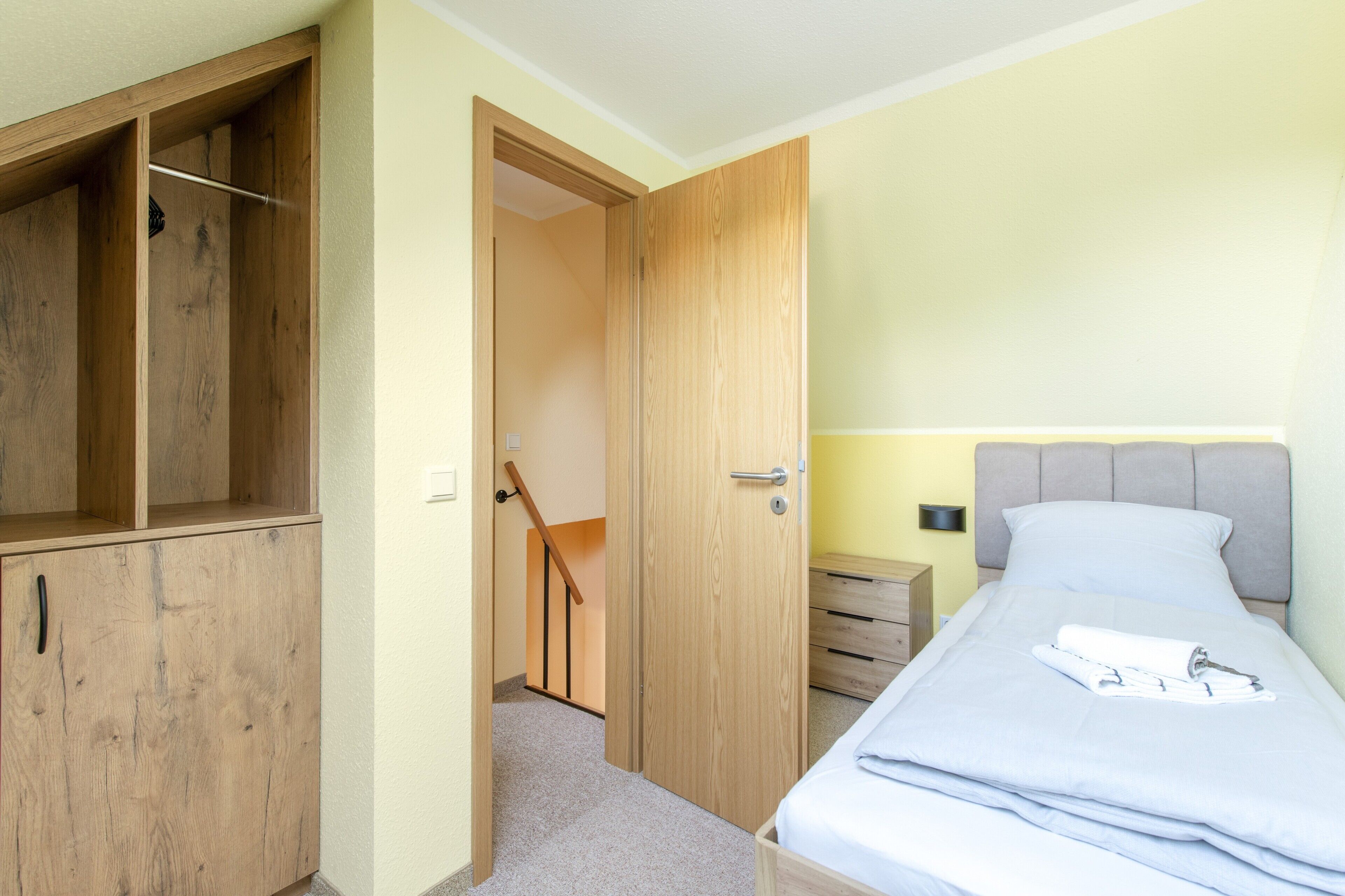 Quarto com cama, guarda-roupa de madeira, mesa de cabeceira e porta para outro cômodo.