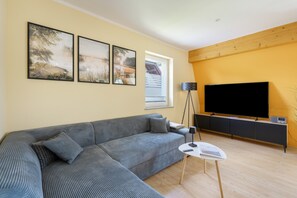 Een moderne woonkamer met een grijze modulaire bank, een flatscreen televisie op een houten dressoir en drie lijstwerken met landschapsprenten aan de muur.