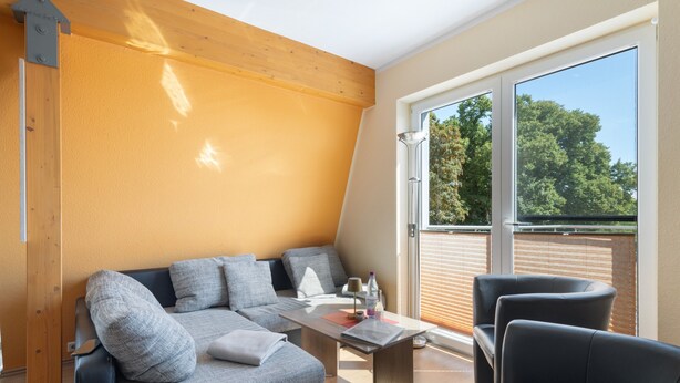 Ein modernes Wohnzimmer mit einer Couch, einem Tisch, Stühlen, einem großen Fenster mit Blick auf Bäume und einer Holzbalkendecke.
