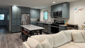 A modern kitchen with grey cabinets, a stainless steel refrigerator, a microwave, and a dining area with stools. -  "Last Light", This Perfect 3-bedroom, 2 bath cottage in quaint GOTL. (Geneva)