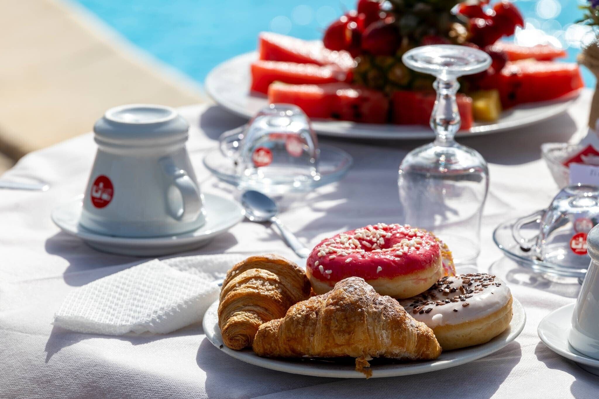 Ein Tisch mit Croissants, einem Donut und einer Wassermelone, im Hintergrund ein Schwimmbecken.