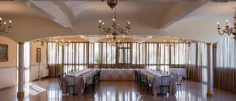 Un amplio salón de banquetes con una larga mesa dispuesta para un evento formal, candelabros ornamentados y grandes ventanales con cortinas.