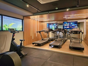 A gym with treadmills, a stationary bike, and a TV screen displaying workout videos. - Beach Haus By Hosppedar (João Pessoa)