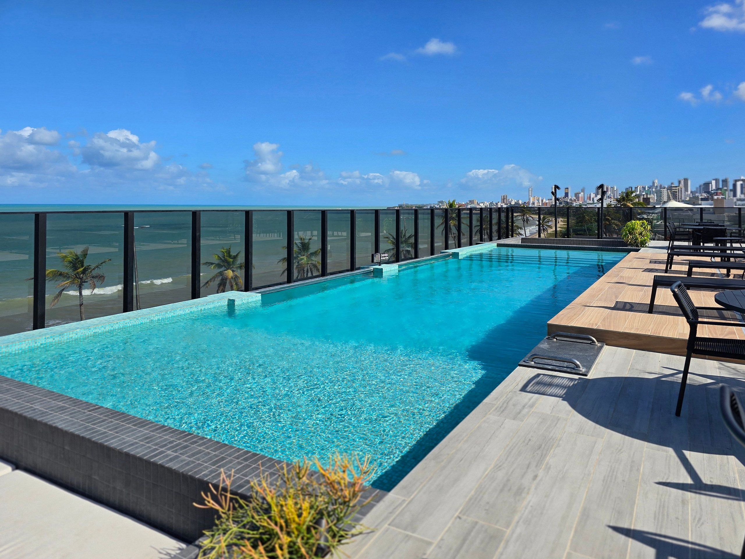 Una piscina infinita en la azotea con vistas a la ciudad, rodeada de sillones de descanso y una terraza de madera.