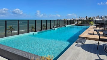 Una piscina infinita en la azotea con vistas a la ciudad, rodeada de sillones de descanso y una terraza de madera.