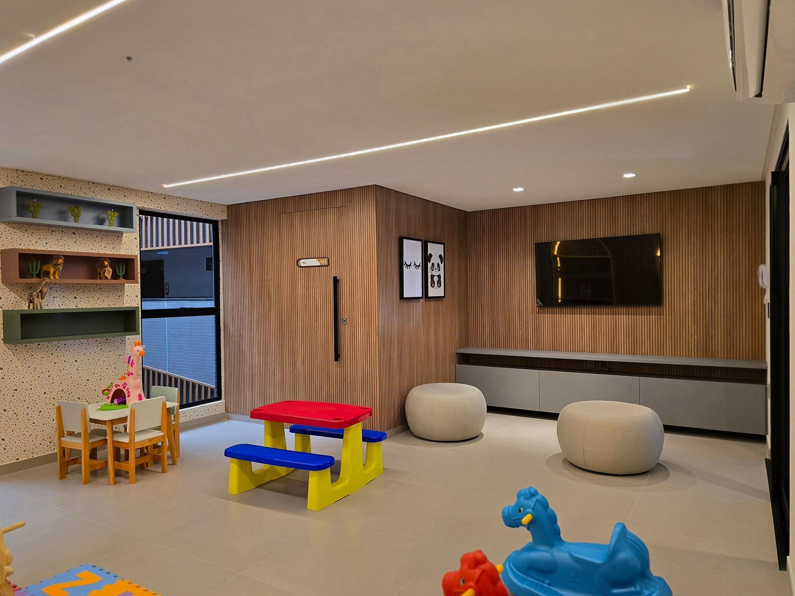 Uma área de recreação infantil moderna, com mesa, cadeiras, um banco e uma TV fixada na parede.