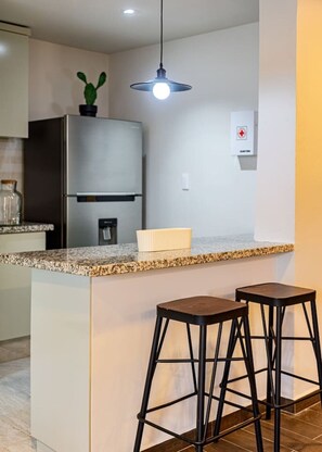 A modern kitchen with a stainless steel refrigerator, a granite countertop, two black stools, and a pendant light. - Group Friendly | Private Terrace & BBQ in Roma Sur (Mexico City)