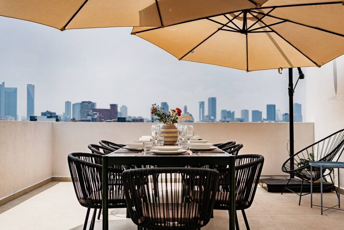 A rooftop dining area with a table set for four, surrounded by black chairs, under a large beige umbrella.