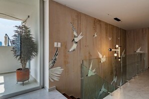Ein modernes Interieur mit einer großen Topfpflanze, einer Wand mit Vogelgemälde und einem Glasgeländer mit Spiegelung.