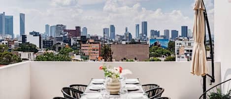 A rooftop dining area with a city skyline view, a table set for four, and a single chair visible.