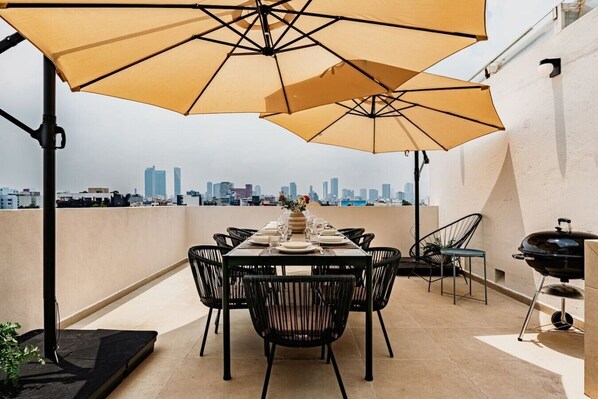 Eine Dachterrasse mit Esstisch, Gartenmöbeln, einem Grill und großen Sonnenschirmen, die Schatten spenden.