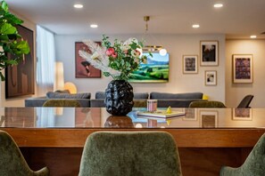 Een moderne woonkamer met een eettafel met glazen blad, groene stoelen, een zwarte vaas met bloemen, een bank en een televisie aan de muur.