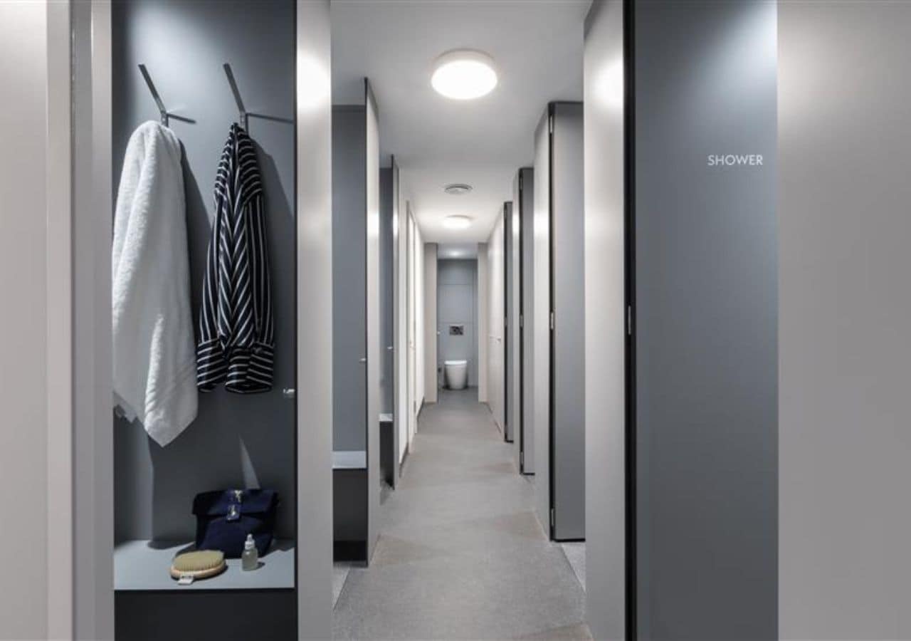 A hotel corridor with a shower area on the right, a bench with a handbag and a bottle, and towels hanging on hooks.