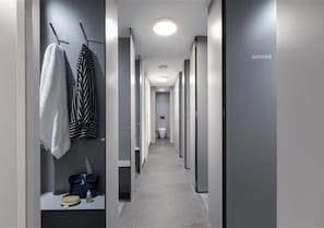A hotel corridor with a shower area on the right, a bench with a handbag and a bottle, and towels hanging on hooks.