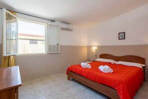 Une chambre à coucher comprenant un lit, une table de chevet, une fenêtre avec des rideaux, un appareil de climatisation et un luminaire fixé au mur.