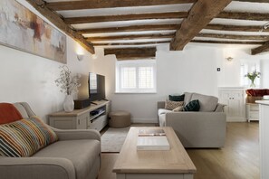 A living room with a television, sofa, and coffee table. There is a window with blinds, a vase with flowers, and a painting on the wall. - The Mews Hideaway -  a cottage that sleeps 4 guests  in 2 bedrooms (Fairford)