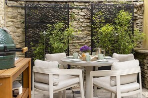 A round table with four white chairs, a decorative wall with climbing plants, and a rustic stone wall in the background. - The Mews Hideaway -  a cottage that sleeps 4 guests  in 2 bedrooms (Fairford)